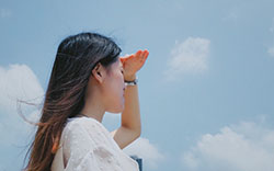 A woman, seen from behind, stands and looks away towards a blue sky.


